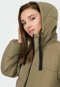 Maslinasto zeleni kaput s podstavom i kapuljačom, koji ima prednji patent zatvarač, crne izvlačne vrpce i kviltanu teksturu za izolaciju.