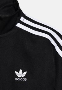 Černý sportovní svetr s bílým třemi pruhy na ramenou a vyšitým logem Adidas na hrudi. Hladký povrch.