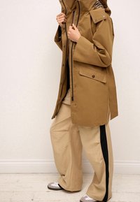 Bruine waterdichte jas met een grote capuchon, voorzakken en zwarte accenten, gecombineerd met beige wijde broek met zwarte zijkanten.