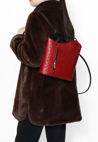 Rode leren rugzak met een gestructureerd oppervlak, voorzien van een ritssluiting en een zwarte band. Draagt over een bruine, pluizige jas.