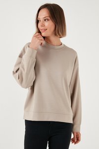 Noor naine lühikeste pruunide juustega, kellel on seljas beež pikkade varrukatega dressipluus ja mustad püksid, vaadates veidi vasakule, käsi lõua lähedal.