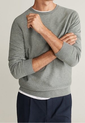 Mand iført en lysegrå langærmet sweater over en hvid skjorte, med navy blå bukser, arme krydset over brystet.
