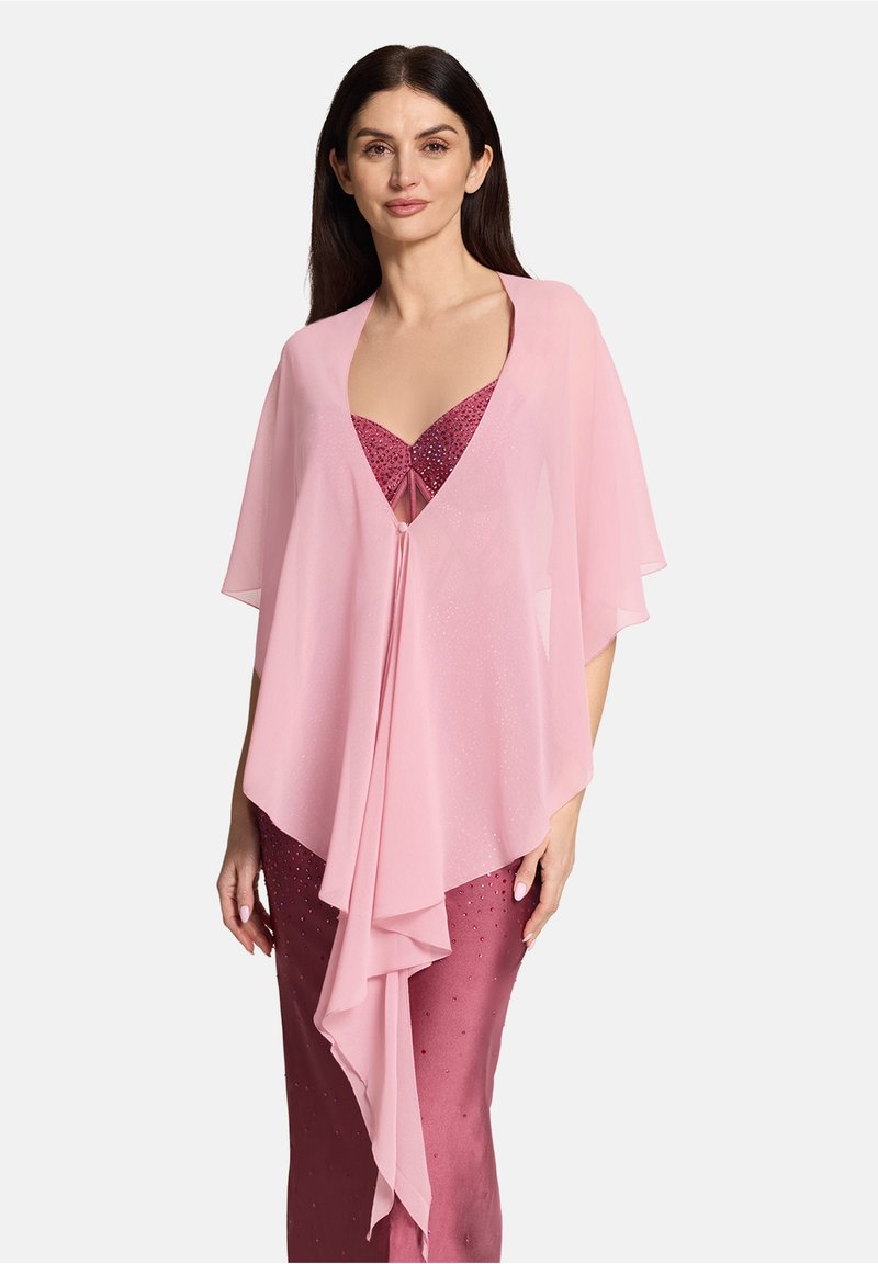 Frau trägt einen durchsichtigen, blassrosa Umhang über einem ärmellosen, tief rosa Pailletten-Kleid und steht vor einem schlichten hellen Hintergrund.