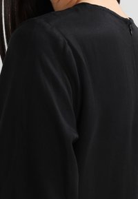 Dos d'une personne portant un vêtement noir à manches longues avec une fermeture éclair au niveau du col et de longs cheveux noirs tombant sur une épaule.