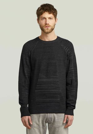 Maglione nero lavorato a maglia con motivi testurizzati, scollatura rotonda e maniche raglan, indossato sopra pantaloni grigi chiari. Design semplice e moderno.