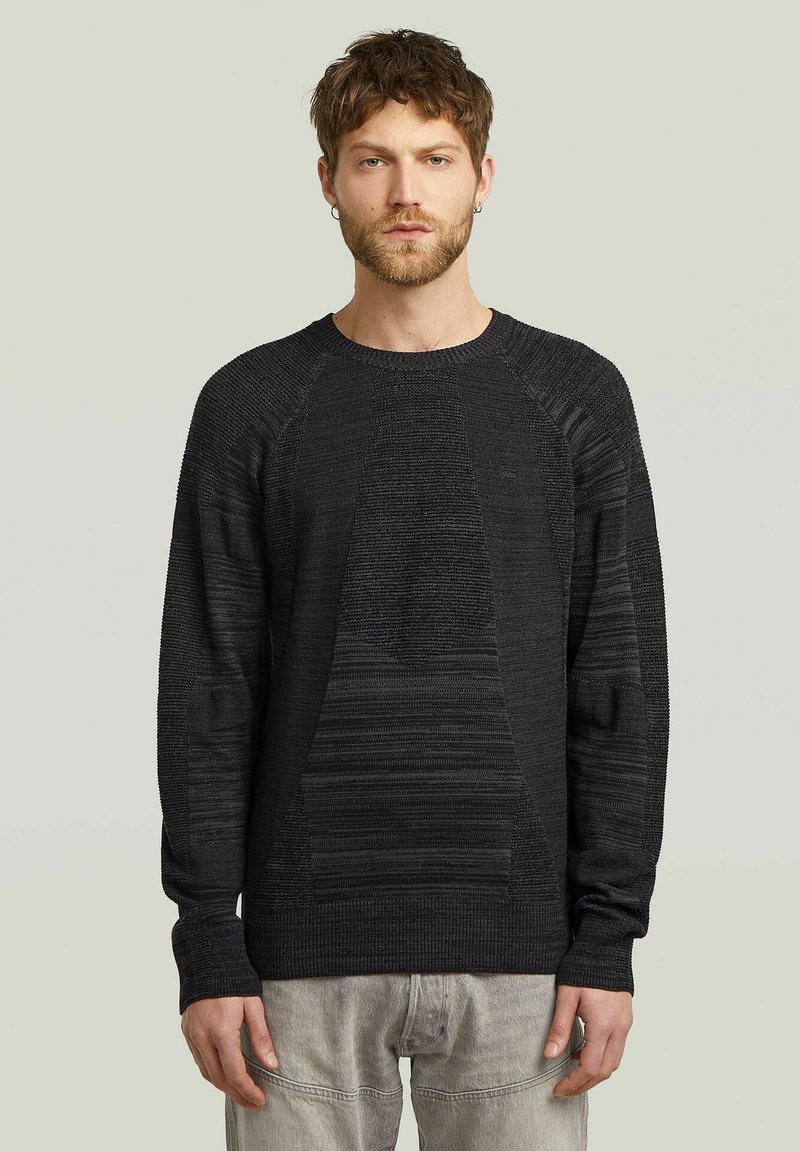 Pull en tricot noir avec des motifs texturés, col rond et manches raglan, porté sur un pantalon gris clair. Design simple et moderne.