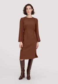 Braunes knielanges Kleid mit rundem Halsausschnitt, puffärmel und seitlichem Schlitz, kombiniert mit knielangen braunen Lederstiefeln. Glatte Textur.