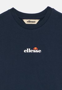 Mørkeblå bomull T-skjorte med rund hals. Har en hvit logo med rød og oransje aksent over teksten "ellesse" på forsiden.