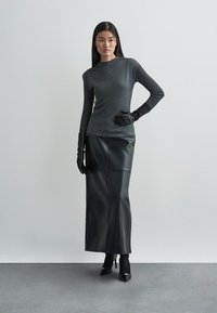 Szürke, hosszú ujjú felső texturált felülettel, fekete bőrmidiruhával, fekete bőrkesztyűvel és hegyes orrú magas sarkú cipővel egy szürke padlón.