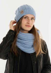 Lichteblauwe gebreide muts met ribbeltextuur en een label. Gecombineerd met een bijpassende lichtblauwe chunky gebreide infinity-sjaal. Zwarte jas.