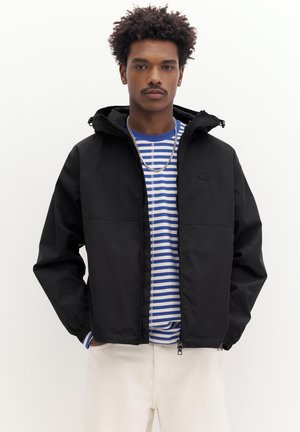 Ung mand iført sort hættetrøje over en blå- og hvidstribet skjorte med hvide bukser, stående foran en ensfarvet baggrund.