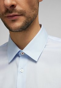 Light blue, textured dress shirt with a pointed collar, featuring white buttons with red accents. Close-up view emphasizes collar and fabric.