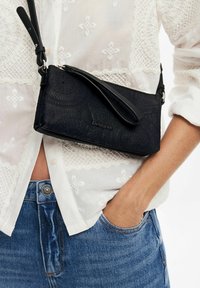Pochette noire texturée avec une bandoulière, ornée d'un motif embossé et d'un détail de logo, posée contre une chemise blanche brodée.