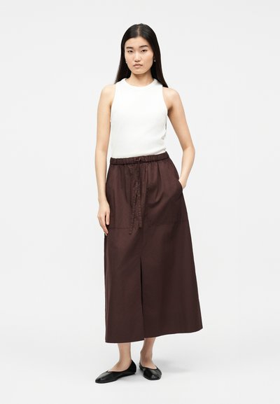 Žena s dlhými tmavými vlasmi, oblečená v bezrukávovom bielom topu, hnedej sukni do členkov so sťahovacím šnúrkou a čiernych šmykľavých topánkach, stojaca na jednoliatom pozadí.