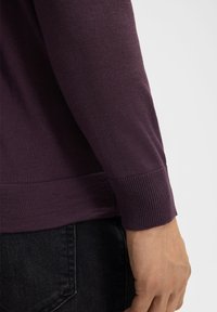 Langanhaltendes lila Strickoberteil mit gerippten Bündchen und Saum. Glatte Textur, figurbetonte Form und minimale Nähdetails am Saum.