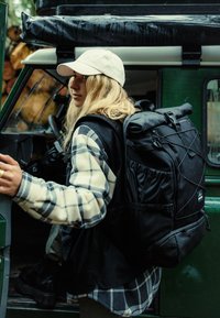 Egy nő fehér sapkában és kockás plédben, nagy fekete hátizsákot cipel, éppen kinyit egy zöld jármű ajtaját szabadtéri környezetben.