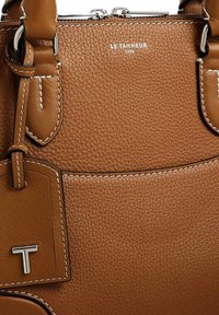 Borsa a tracolla in pelle marrone con superficie testurizzata, cuciture bianche a contrasto e un etichetta metallica con la lettera "T". Chiusura a zip sulla parte superiore.