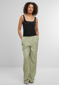 Top negro sin mangas con escote cuadrado combinado con pantalones amplios plisados de color verde claro. La modelo tiene el cabello rizado y las manos en los bolsillos.