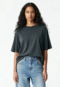 Femeie cu părul lung și creț, purtând un tricou larg de culoare gri închis, introdus în blugi deschiși la culare cu talie înaltă, pe un fundal simplu.