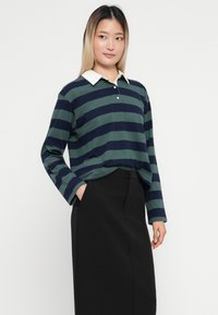 Polo à manches longues à rayures vert foncé et bleu marine, avec un col blanc, une patte de bouton et une coupe décontractée. Porté avec un pantalon noir taille haute.