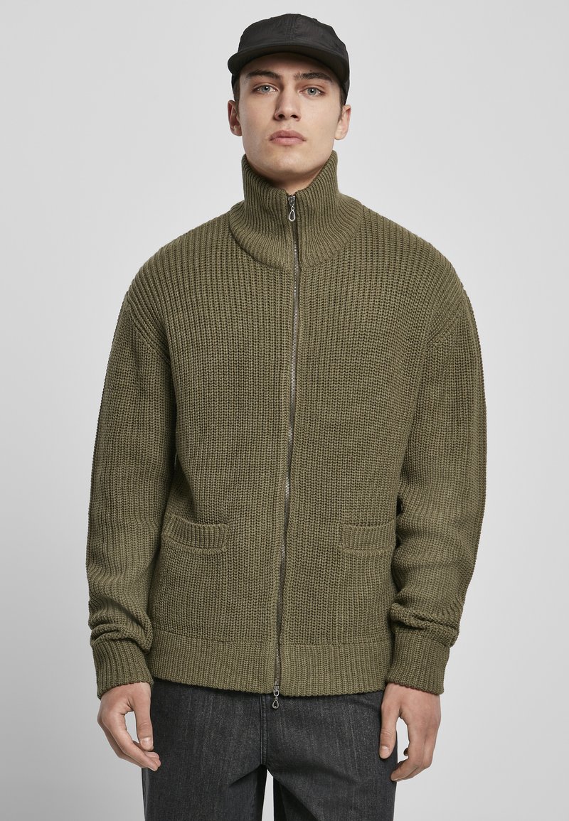 Olivgrön stickad zip-up tröja med hög krage, texturerad tyg, två framfickor och ribbade ärmar. Kombinerad med mörkgrå byxor.