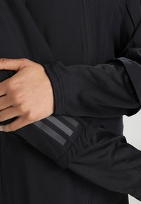 Veste légère noire dotée d'une fermeture éclair sur le devant, d'une texture lisse et de trois rayures réfléchissantes verticales sur la manche.