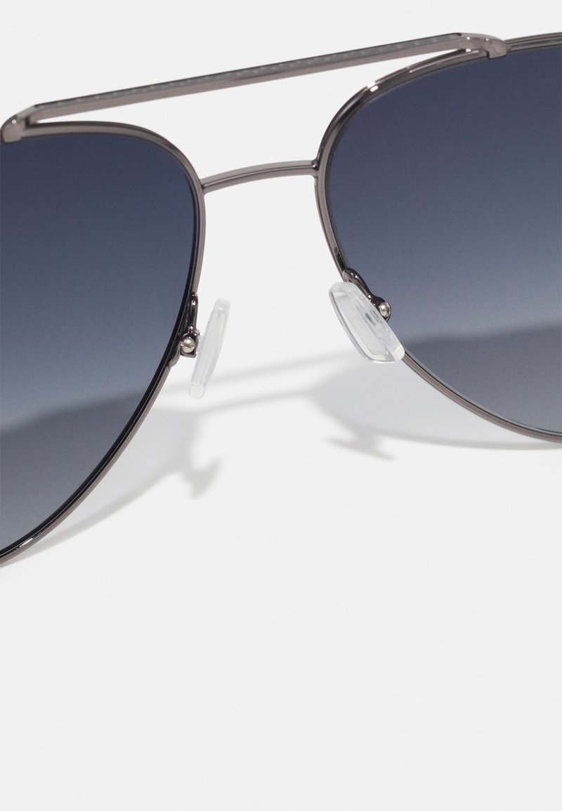 Des lunettes de soleil style aviateur avec des verres dégradés sombres et une fine monture en métal argenté reposant sur une surface blanche.