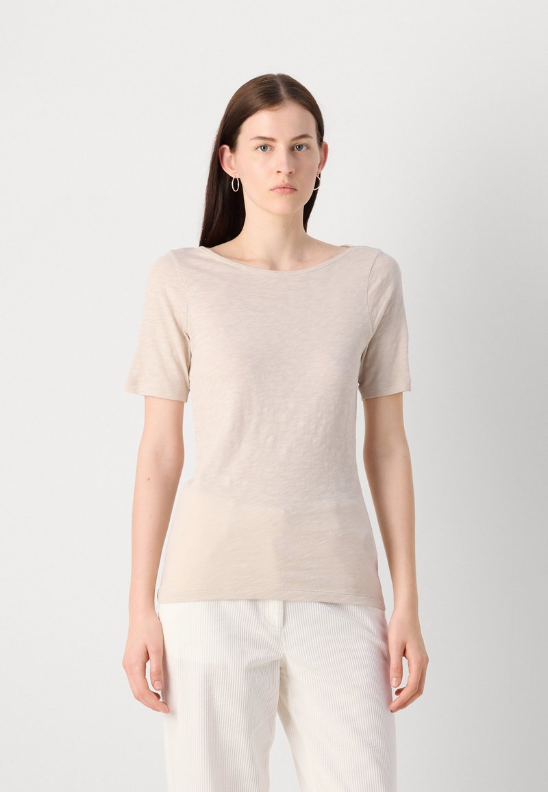 Beige T-Shirt mit kurzen Ärmeln und weiter, runder Ausschnitt, aus einem leichten, strukturierten Stoff. Kombiniert mit weißen gestreiften Hosen.