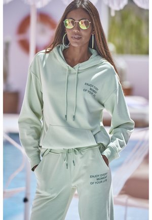 Mint green hoodie and joggers in soft fabric. Features kangaroo pocket and drawstrings, with printed text on both pieces. Sunglasses and large hoop earrings.