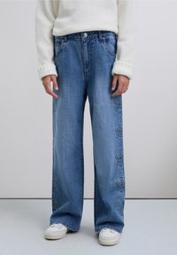 Vaqueros acampanados de mezclilla azul claro con bordado en forma de estrella a lo largo del lateral exterior. Cuenta con cierre clásico de botón y cremallera.