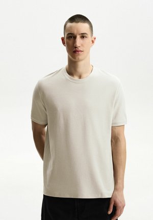 Helles beige T-Shirt mit kurzen Ärmeln aus strukturiertem Stoff. Verfügt über einen runden Halsausschnitt und einen lockeren Schnitt, ohne sichtbare Muster oder Grafiken.