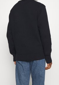 Persona che indossa un maglione nero lavorato a maglia e jeans di denim blu, vista da dietro su uno sfondo bianco semplice.