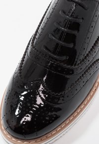 Gros plan d'une chaussure Oxford en cuir verni noir brillant avec des détails brogue et des lacets noirs, présentant un bord de semelle cousu couleur beige.