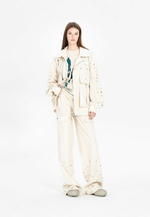 Joven mujer con pantalones y chaqueta cargo color crema con puntos plateados, camiseta gráfica beige y zapatos beige texturizados, de pie sobre fondo blanco.