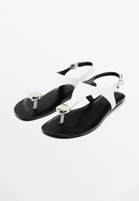 Hvide og sorte sandaler med flad sål, der har et elegant design, krydsende stropper og sølvdetaljer på tæeremmen.