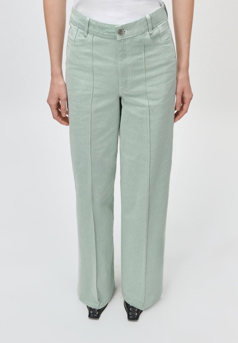 Γυναίκα που φοράει παντελόνι μέντας ψηλόμεσο με φαρδιά μπατζάκια και μπροστινές ραφές, συνδυασμένο με μαύρα παπούτσια μυτερά και λευκό μπλουζάκι μέσα από το παντελόνι.