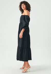 Svart, off-shoulder, lager-på-lager klänning med texturerad tyg. Har puffärmar i lång längd och en flödande silhuett, kombinerad med vita remsandaler.