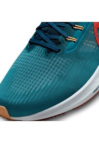 Tyrkysová sportovní obuv má strukturovaný textilní svršek s tmavě modrými tkaničkami, oranžovými detaily a b�ílou mezipodešví s červenou gumovou podešví.