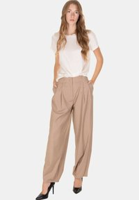 Donna con capelli lunghi e mossi, indossa una maglia bianca a maniche corte, pantaloni beige a vita alta e gamba larga, e tacchi alti neri, con le mani in tasca.