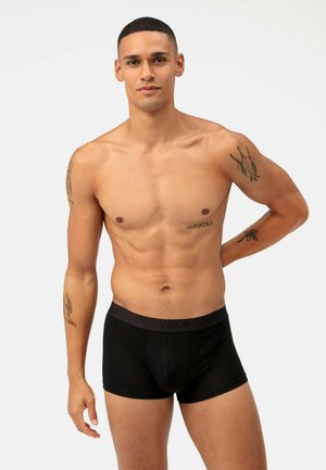 Boxer-briefs noirs en tissu doux avec une texture lisse. Dotés d'une ceinture sombre avec le logo "OLYMP". Un homme se tient debout, les bras détendus.