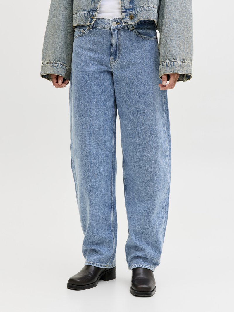 Personne portant un jean large bleu clair, des bottes noires à bout carré et une veste en jean courte par-dessus un haut blanc, debout sur un fond uni.