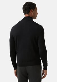 Schwarzer Strickpullover mit hohem Kragen und langen Ärmeln. Glatte Textur. Tailliertes Design mit Details an Schulter und Bündchen. Wird mit grauer Hose getragen.