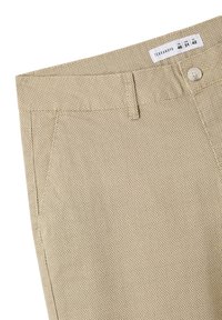 Pantaloni beige intrecciati con un motivo testurizzato, dotati di chiusura con bottone, passanti per cintura e un'etichetta con le informazioni sulla taglia.