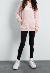 Felpa rosa chiaro con tasca frontale e logo bianco, abbinata a leggings neri con un emblema colorato di lato e sneakers bianche.