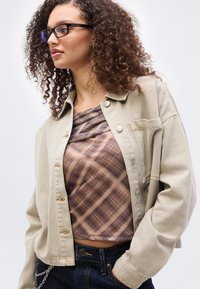 Γυναίκα με σγουρά μαλλιά που φοράει μαύρα γυαλιά, μπεζ μπουφάν, καρό μπλουζάκι και σκούρο τζιν με το χέρι στην τσέπη, κοιτάζοντας προς τα αριστερά.