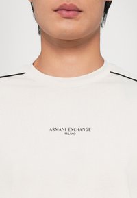 Camiseta de algodón blanca con cuello redondo, costuras de acento negras en los hombros y el logotipo impreso "ARMANI EXCHANGE MILANO" en el centro.