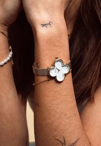 La montre-bracelet en maille dorée présente un cadran blanc en forme de fleur avec des détails dorés, de petits repères horaires et un accent en chaîne.