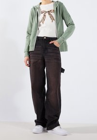 Sudadera verde con cremallera, camiseta blanca con un lazo de estampado de leopardo, vaqueros negros holgados y deportivas blancas. Ajuste casual y relajado.