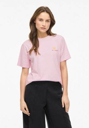 Kvinde iført en lyserød T-shirt med en lille gul sol og teksten "le soleil", parret med højtaljede sorte bukser, står neutralt.