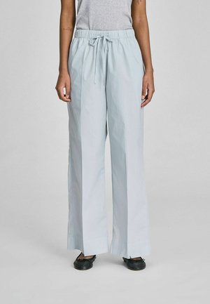 Inimene, kes kannab helesiniseid laia säärega paelaga pükse ja musti baleriinasussid, seisab ühevärvilise hele tausta ees.